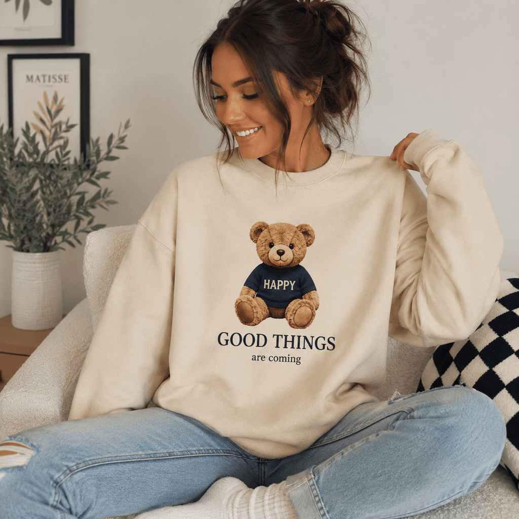Woman wearing a beige sweatshirt with a teddy bear graphic and text, sitting on a couch.